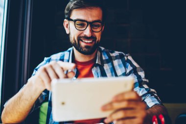 Modern tablet tarihinde yayına izlerken gözlük olumlu sakallı hippi adam bağlı 4 g için Internet. Casual kıyafet online dijital dokunmatik ped cihaz üzerinde sohbet içinde neşeli erkek blogger