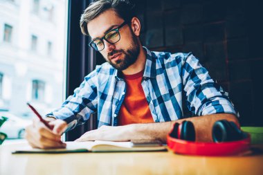 Dalgın sakallı öğrenci ödev Not defteri alanı coworking eğitim aşağı yazma. Akıllı hipster adam ders notları not defterinde kafede sınava hazırlık sırasında yapma gözlük