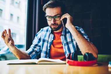 Kızgın genç adam kitap alan coworking içinde vasıl okul sırası oturan smartphone aygıt üzerinde iletişim gözlük sakallı. Telefon görüşmesi sırasında konuşurken rahat kıyafet rahatsız hipster adam giymiş