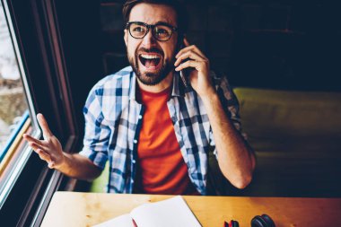 Kızgın sakallı hippi biri üstten görünüm alanı coworking çalışma dijital Smartphone cep telefonu ile telefon görüşmesi sırasında rahat kıyafet bağırarak giymiş. Ağlama ve hareket eden telefon aygıt üzerinde savunarak öfkeli genç adam