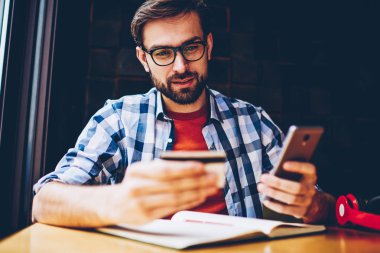 Dalgın casual giyinen hipster adam web sitesinde sipariş yapmak için kartvizit smartphone telefon numarasını çevirme. Yapım para hareket online üstünde hareket eden telefon içinde coworking oturan genç adam