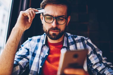 Pensive bearded young man in optical eyeglasses for vision correction holding banking card in hands while making payment online on website using free internet connection on smartphone device clipart