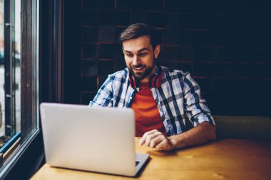 Neşeli sakallı öğrenci komik eğitsel bir Web sitesinde modern netbook aygıt izlerken boyun kulaklık ile kablosuz internet'e bağlı. Haber üstünde laptop blog okumak pozitif genç adam serbest meslek