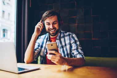 Modern kulaklık dinlemek için neşeli sakallı hippi adam şarkı müzik uygulaması seçme smartphone için bluetooth ile bağlantılı. Olumlu erkek blogger cep çalar ses tadını çıkarırken sohbet