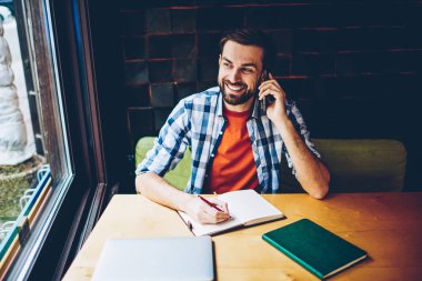 Kafede masada oturan Not defteri ödev yaparken smartphone aygıt üzerinde arama başarılı sakallı öğrenci. Not defterinizdeki notlar aşağı yazarken telefonla konuşurken neşeli hipster adam