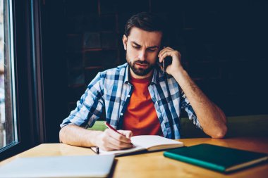 Konsantre hipster adam coworking masada oturan smartphone üzerinde konuşma sırasında duydum Not Defteri metin bilgileri yazma rahat forması giymiş. Cep üzerinde calling düşünceli öğrenci