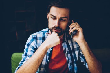 Coworking içinde oturan Smartphone cep telefonu ile telefon görüşmesi sırasında operatör ve değişim tarife planı hakkında düşünme dinleme telefon önerisi düşünceli sakallı genç adam. Sakallı hippi adam cep üzerinde bahsediyoruz