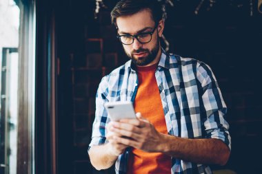 Dalgın sakallı hippi blogger gelen bildirim okuma ve ağlar Smartphone cep telefonu ile sohbet bağlı 4 g için Internet. Genç adam cep telefonu online video izlerken rahat gömlek giymiş