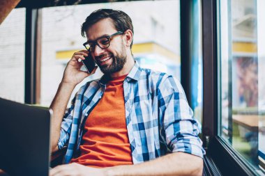 Happy sakallı genç adam gözlük smartphone yazılım yolu ile radyo 4 g modern dizüstü bilgisayarda güncelleştirilirken iletişim internet. Netbook hücresel oturma konuşurken olumlu hipster adam