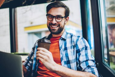 Yarım uzunluk portre mutlu yakışıklı el ve kameraya gülümseyen gitmek için kahve tutan gözlük öğrenci sakallı. Uzay coworking dinlenme içecekler ile olumlu erkek blogger