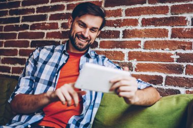 Mutlu sakallı genç adam modern Dokunmatik ped cihaz üzerinde Web sitesinde komik Haberler okuma rahat gömlek giymiş. Neşeli hipster blogger yayın 4 g internet bağlantısı üzerinden dijital tablet İndiriyor