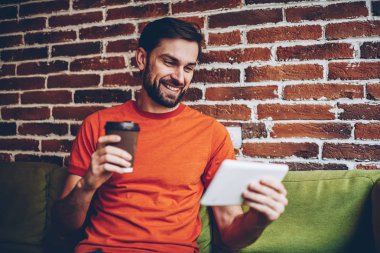Gülümseyen sakallı erkek bogger kahve paket servisi olan restoran izlerken komik canlı stream sosyal ağ üzerinden Internet dijital tablet, e-posta dokunmatik yüzey üzerinde iyi haber okumak pozitif genç adam bağlı 4 g ile