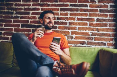 Pozitif sakallı bogger kahve ile gitmek el gülümseyen ve uzak alan coworking içinde kanepede oturan 4 g internet bağlantısı ile smartphone aygıtında mobil uygulama yüklenirken arıyorsunuz