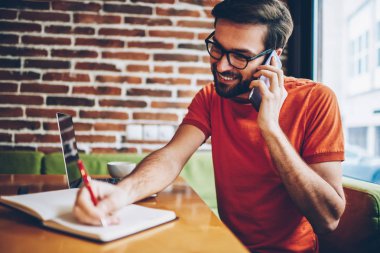 Neşeli smartphone aygıt üzerinde arama ve metin bilgileri dizüstü cihaz alan coworking içinde oturan Not Defteri'nde yazma genç adam sakallı. Telefonla konuşurken ve notlar olumlu hipster adam