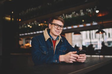 Yarım uzunluk portre denim ceket şık kahve dükkanında dinlenme 4g internet bağlantısı ile Smartphone cep telefonu ile sosyal ağlarda profil güncelleştirilirken kameraya gülümseyen başarılı erkek Blogger