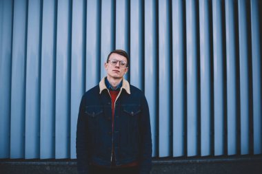 Şık hipster bogger yarım uzunluk portre üzerinde promosyon arka plan açık havada poz denim ceket giymiş. Kentsel ortamda kamera ayakta bakarak tasarım giyim harika genç adam