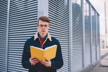 Dalgın zeki öğrenci edebiyat kitap ayakta Caddesi üzerinde kentsel ortamda okuma rahat denim ceket giymiş. Hobi ücretsiz vakit elinde yenilikçi genç adam ders kitabı ile konsantre