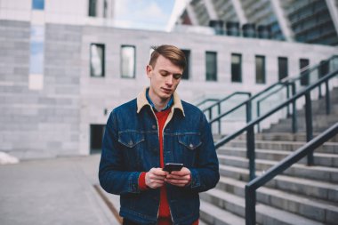 Dalgın genç blogger posta ile bildirim Smartphone cep telefonu ile kontrol bağlı 4 g için Internet açık havada geziniyor. Hipster adam sokakta yürürken telefonu uygulaması güncelleştiriliyor denim ceket