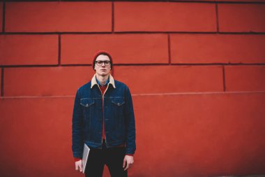 Gözlük kamera dizüstü bilgisayar elinde tutarken arıyorum ciddi rahat giyinmiş genç adam portresi. Şık denim ceket açık havada promosyon arka plan üzerinde duran hipster adam