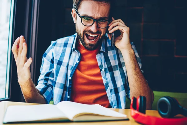 Furious bearded hipster guy shouting during phone conversation on digital smartphone.Angry young man arguing and solving dispute calling on modern cellular device sitting at desktop with textbook