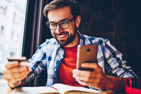 Cheerful bearded student with banking card in hands making payment on website on smartphone via internet connection.Positive young man looking at business card holding phone for money transaction