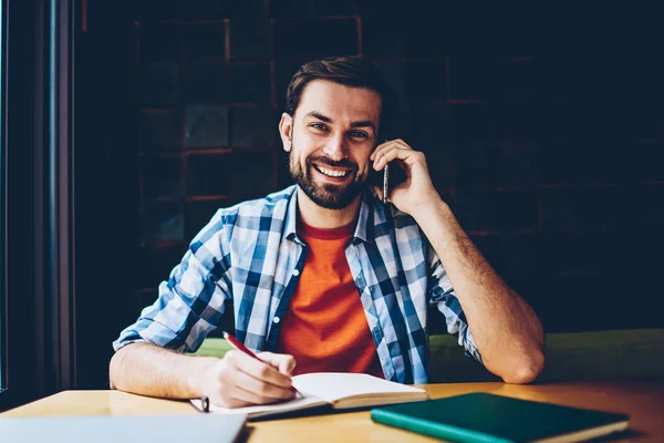 一个成功的胡子学生的肖像微笑着在相机 而呼吁智能手机和笔记在记事本坐在 Coworking 积极的时髦家伙沟通电话设备 — 图库照片
