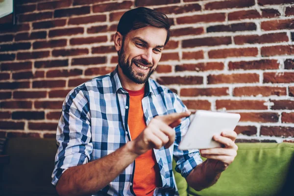 Successful bearded young man dressed in casual shirt typing text information chatting online in social networks on modern tablet via free 4G internet.Positive hipster blogger updating profile