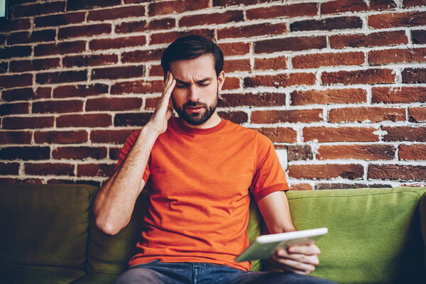 Stressed bearded man dressed in orange t-shirt has migraine.Unhappy hipster guy has headache while sitting on sofa with modern tablet device in coworking space.Exhausted male with touch pad