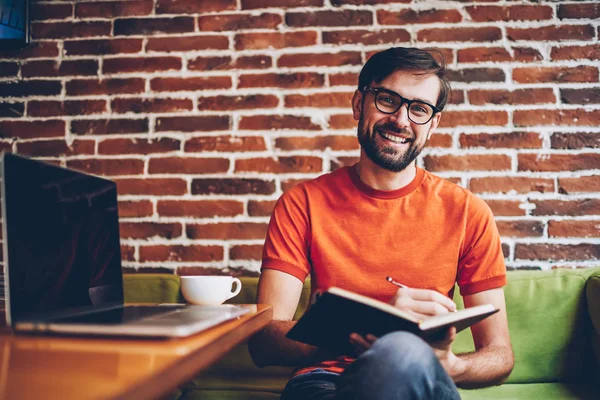 Half length portrait of successful bearded young man in eyewear smiling at camera while writing down text information in notepad to plan working process sitting at modern netbook in coworking space