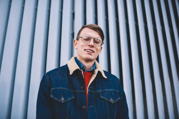 Half length portrait of cheerful young student dressed in fashionable denim apparel laughing at camera.Positive hipster guy in optical eyeglasses for vision correction posing in urban setting