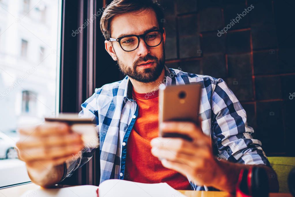 Bearded young man in eyeglasses for vision correction holding business card in hands and dialing phone number on smartphone to call.Pensive handsome male blogger searching information on telephone
