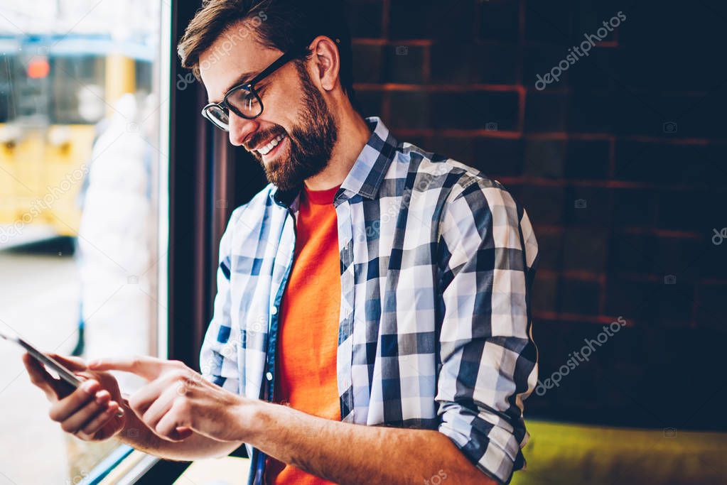 Joyful hipster young man in eyewear typing text message on smartphone device.Positive student reading funny notification and chatting in social networks on telephone via free 4G internet connection