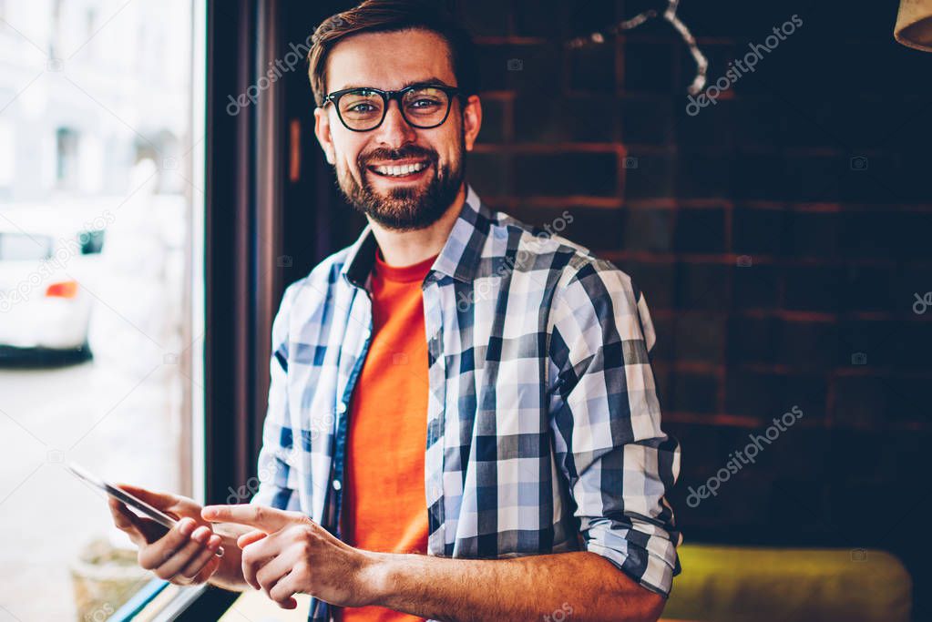 Portrait of successful bearded hipster blogger in eyeglasses smiling at camera while updating profile on smartphone via 4G internet.Happy young man in casual stylish shirt holding mobile phone in hand