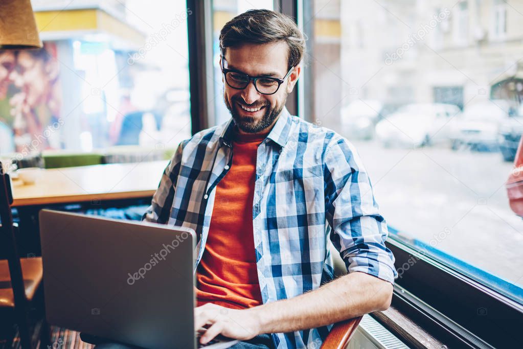 Cheerful bearded hipster guy laughing and chatting online with friends in social networks on laptop connected to wireless 4G internet.Positive male blogger reading funny news on netbook in coworking