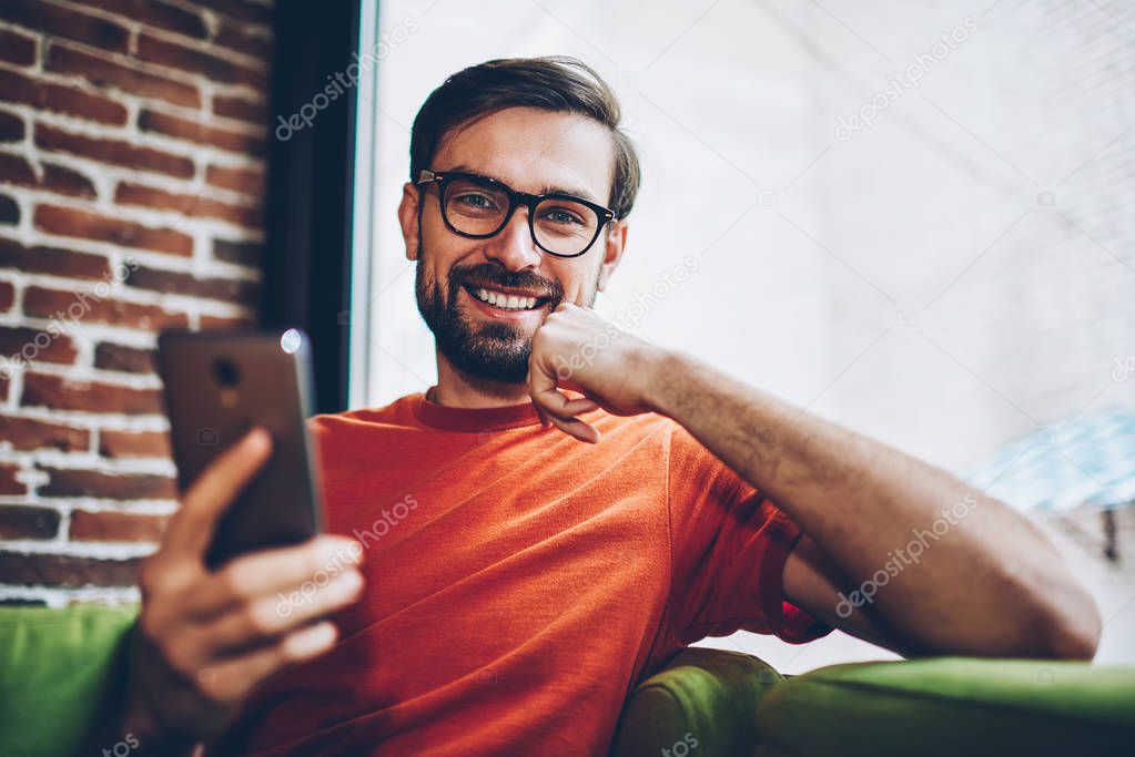 Portrait of successful bearded male blogger laughing at camera while downloading app on smartphone device via free 4G internet connection.Positive hipster guy making payment online on cellular