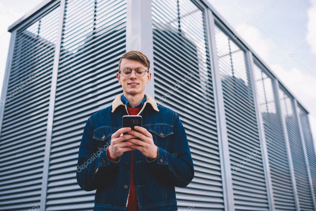 Successful hipster blogger laughing during friendly messaging in social networks on smartphone connected to 4 internet.Positive young man updating profile on telephone standing in urban setting