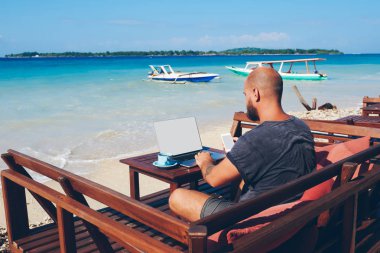 İşadamı rahat kıyafet online netbook ve kablosuz internet, rezervasyon rezervasyon sahilde oturan App kontrol ederken erkek bin yıllık holding cep telefonu kullanarak adada tatil sırasında çalışma