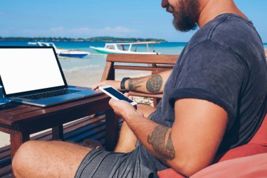 Mobil Bankacılık denge kontrol ve okyanus plaj, Erkek freelancer teknolojisini kullanarak uzak iş sırasında oturan işlem yapmadan telefon tutarak adamın kırpılmış görüntü senkronize cep telefonu ve netboo