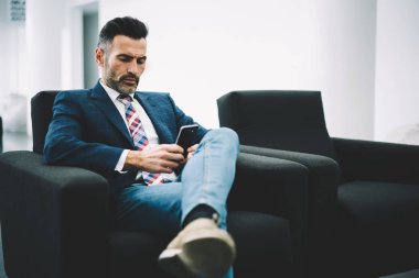 Dalgın olgun girişimci giyinmiş formal giyim finansal haber yolu ile 4 g Smartphone cep telefonu ile internet sitesinde okumak içinde ofis iç koltukta oturan internet. Gurur CEO'su posta kontrol konsantre
