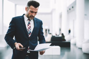 Dalgın ciddi girişimci dikkatle finansal raporlar ofiste ayakta okuma takım elbise giymiş. Olgun işadamı smartphone ile kağıt belgeleri kontrol elinde konsantre