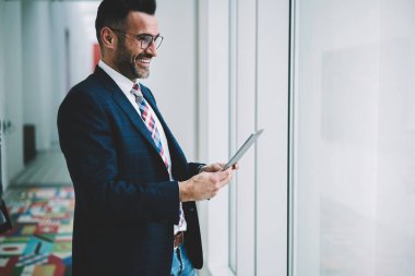 Formal giyim pencereden dışarı bakarken posta kontrol etmek için dijital tablet holding neşeli müreffeh işadamı 50 yaşında giymiş. Pozitif olgun gurur CEO'su para hareket sırasında gülüyor