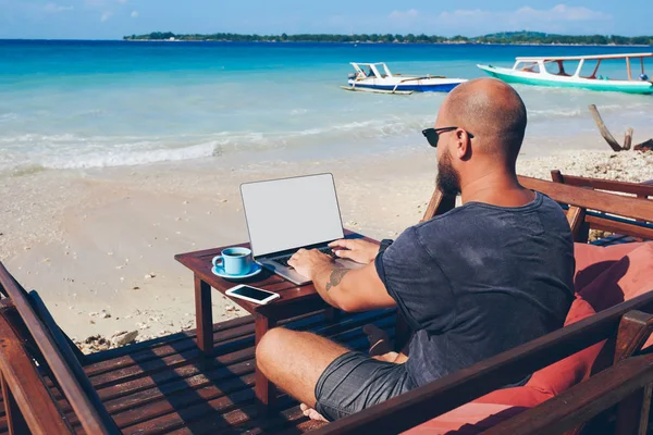 Back view of young man sitting with laptop computer on sea shore working online during vacations, professional male freelancer keyboarding on netbook with mock up screen enjoying summer trip