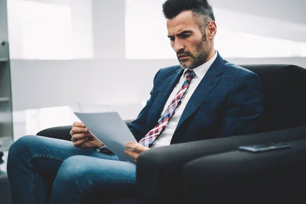Concentrated experienced mature proud ceo dressed in formal wear reading accounting reports on papers sitting on armchair in office.Pensive businessman 50 years old checking financial information