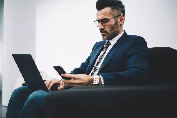 Confident executive manager in formal wear concentrated on work at modern netbook sitting in office.Pensive entrepreneur middle aged with smartphone in hand checking banking account on laptop computer