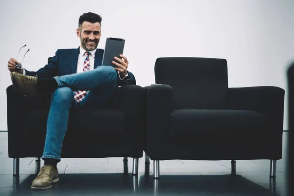 Cheerful successful proud ceo dressed in formal wear watching funny videos on touch pad connected to 4G internet sitting on armchair.Positive mature entrepreneur laughing while reading news on tablet