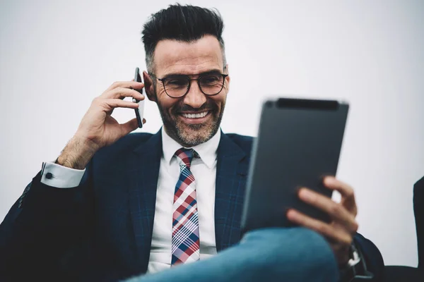 Cheerful mature proud ceo 50 years old dressed in formal wear read funny information on digital touch pad and calling to business partner on smartphone.Happy financial director talking on mobile phone