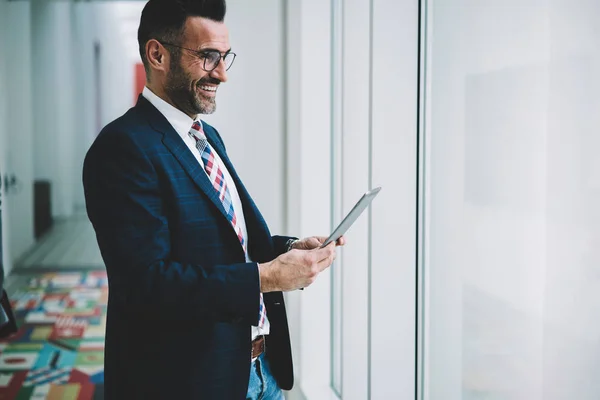 Cheerful prosperous businessman 50 years old dressed in formal wear holding digital tablet to check mail while looking out of window.Positive mature proud ceo laughing during money transaction