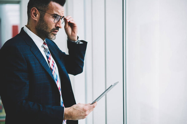Side view of thoughtful confident proud ceo 50 years old looking out of window while updating website with financial news on tablet connected to 4G internet.Pondering successful trader with touch pad