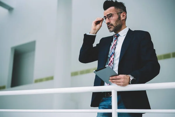 Thoughtful confident entrepreneur 50 years old holding digital tablet in hands.Pondering mature businessman dressed in formal wear thinking on notification received on modern touch pad device