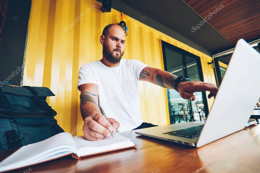 Clever caucasian male student copying information from web page to notebook doing homework using laptop computer in college campus, millennial man pointing on netbook monitor and making notes